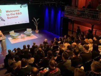 Picture of the opening of the IDFA DocLab R&D Summit taken from the balcony showing a theatre full of people in the audience listening to a women on stage speaking.