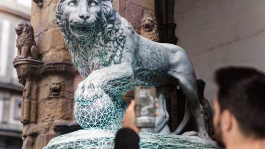 Picture with a man in the foreground holding a cell phone that's pointed at a lion statue in the city that he is volumetrically scanning with Niantic's Lightship AR APIs