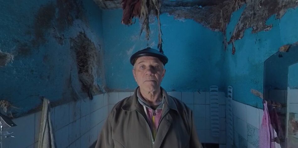 Older Ukrainian man standing within a bombed out bathroom while staring within a 360 video camera.