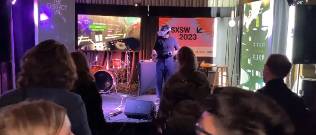 Picture of Dennis Lisk on stage at SXSW wearing a VR headset and controlling virtual knobs as he plays a live DJ set using The District VR software.